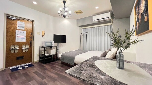 Habitación de hotel con cama, escritorio con televisor de pantalla plana, microondas, zapatero y puerta con letrero 'Host'.