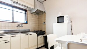 A compact kitchen with white cabinets, a gas stove, a microwave, and a small dining area with a white table and chair. - Ikebukuro Yamase Building GR1 (Tokyo)