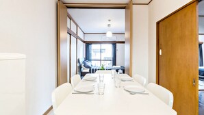 A dining area with a white table set for six, a wooden door on the right, and a view into a room with a bed and window. - Ikebukuro Yamase Building GR1 (Tokyo)