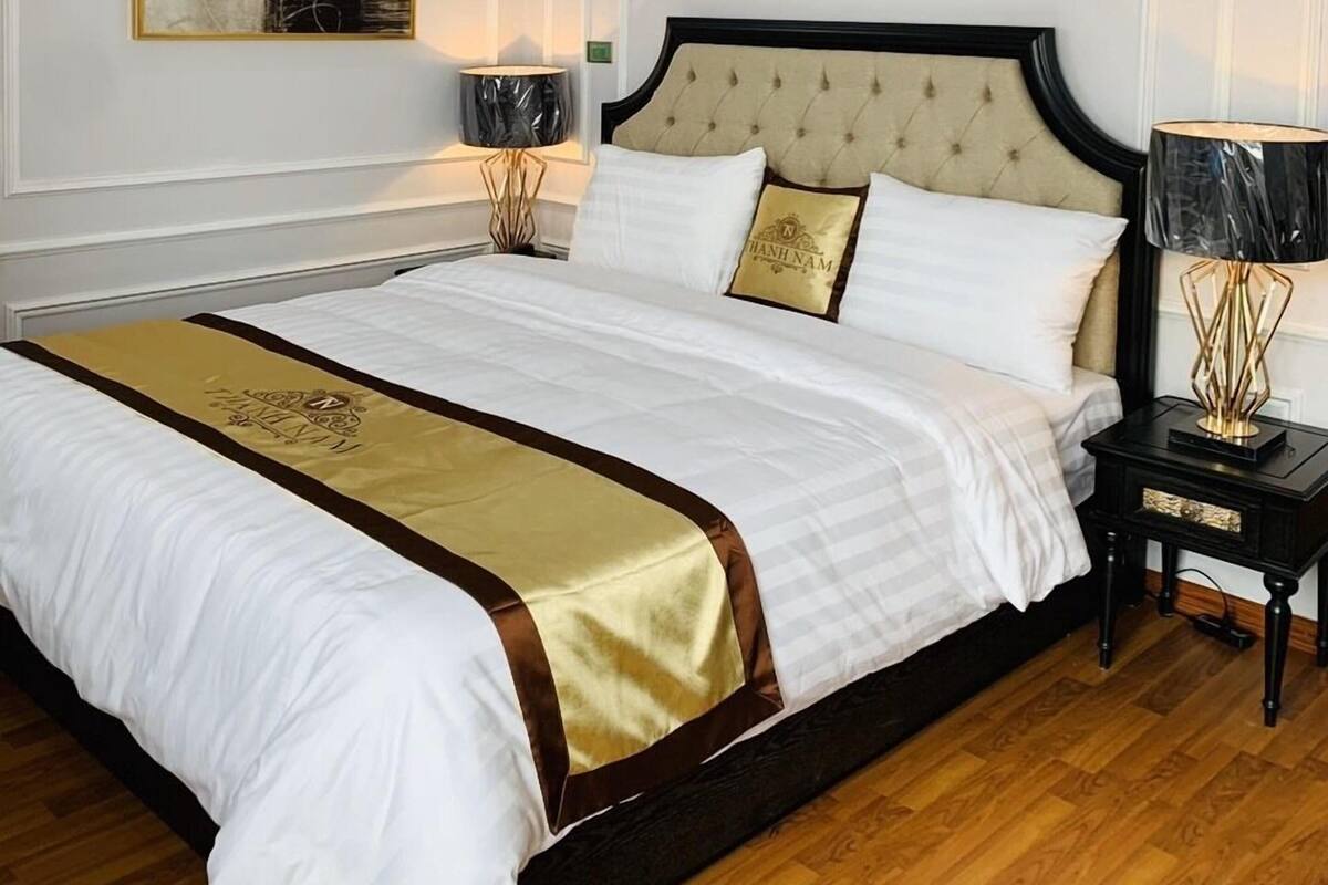 A neatly made bed with a gold accent, a bedside table with a lamp, and a framed abstract painting on the wall.