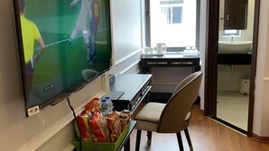 Quarto de hotel com televisão de tela plana exibindo uma partida esportiva, uma escrivaninha com cadeira, uma mesinha com petiscos e vista para o exterior através de uma janela.