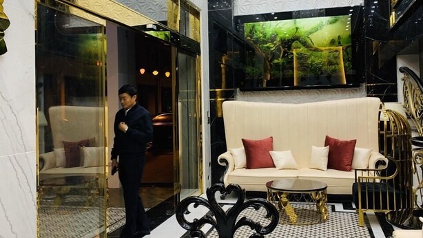 Um hall de entrada luxuoso com um sofá branco, almofadas vermelhas, uma mesa de centro preta e dourada, e uma tela grande exibindo uma cena da natureza.