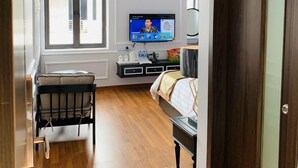 Quarto de hotel com chão de madeira, uma cama, televisão na parede, uma pequena mesa com cadeira e uma janela com persianas.