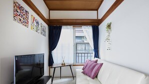 A modern living room with a flat-screen TV, a white sofa with purple cushions, a small table with a vase and candles, and a balcony with blue curtains. - RESIDENTIAL HOTEL DAIICHI-H SHIN-OKUBO Room.A (Tokyo)