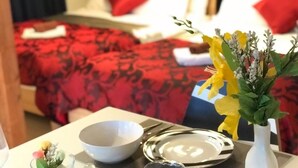 A hotel room with a bed covered in red bedding, a white pillow, and a red pillow on the bed. There is a table with a white bowl, a silver plate, and a vase with flowers. - RESIDENTIAL HOTEL DAIICHI-H SHIN-OKUBO Room.A (Tokyo)