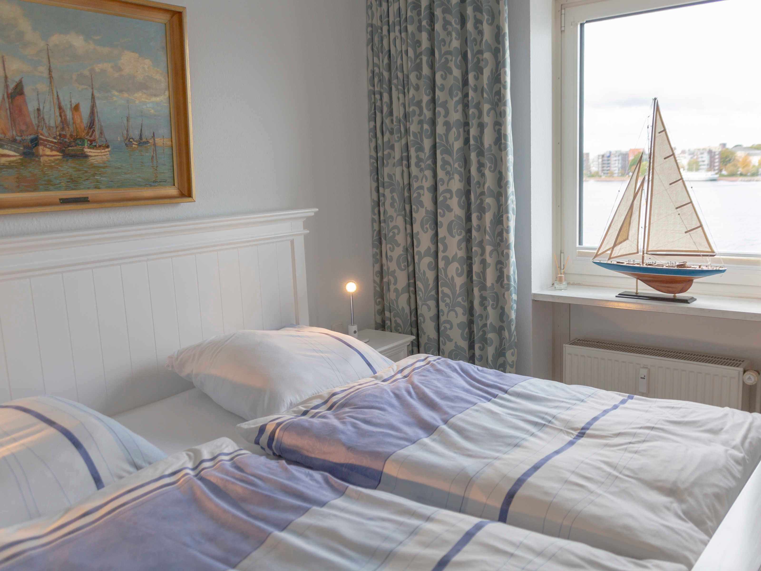 A bedroom with two beds, a painting of sailboats, a window with a sailboat model, and a nightstand with a lamp.