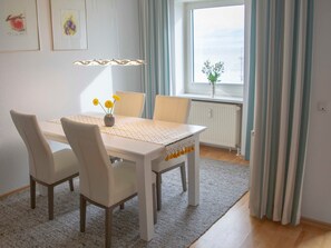 Comedor con una mesa blanca, cuatro sillas, un jarrón con flores amarillas y una ventana con cortinas.