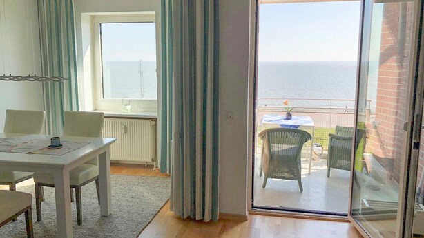 Une pièce comprenant une table à manger, des chaises, un radiateur, une fenêtre et une porte-fenêtre coulissante donnant sur un balcon avec vue sur la mer.