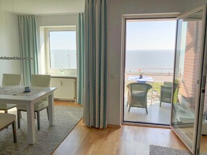 A room with a dining table, chairs, a radiator, a window, and a sliding glass door leading to a balcony with a view of the sea. - 107-2 Neptun - 107-2 Ferienwohnung Neptun (Wilhelmshaven)