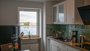 Une cuisine compacte avec des meubles blancs, un réfrigérateur noir, une fenêtre donnant sur des bâtiments et l’eau, un évier et une machine à café.