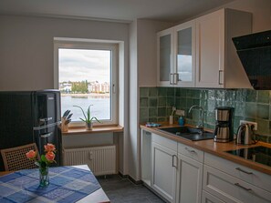 Una cocina compacta con armarios blancos, un refrigerador negro, una ventana con vista a edificios y agua, un lavamanos y una cafetera.