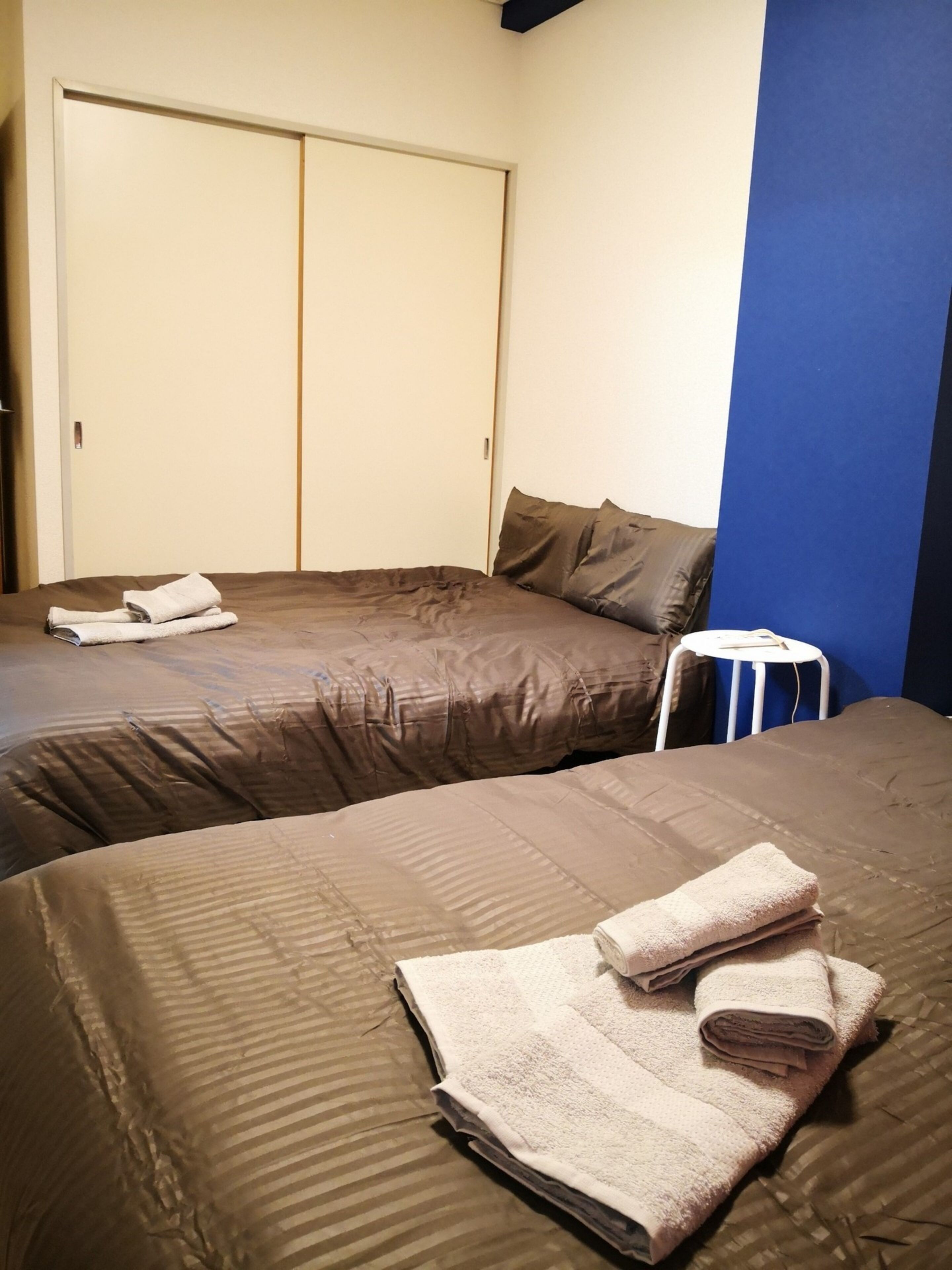 Habitación de hotel con dos camas, cada una con ropa de cama y almohadas marrones, y un par de toallas sobre cada cama. En la esquina hay una silla blanca y una mesita redonda pequeña.