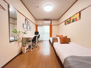 A hotel room with a bed, a desk, a chair, a mirror, a plant, and a painting on the wall. - RESIDENTIAL HOTEL YAMATO-H Shimokitazawa 202 (Tokyo)