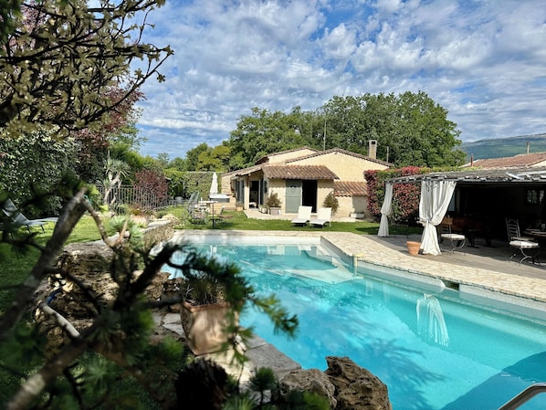A pool with a stone border, a house with a tiled roof, and a garden with various plants.