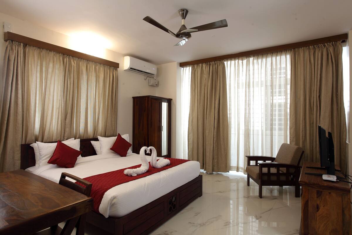 Quarto de hotel com uma cama grande, uma escrivaninha de madeira, uma cadeira, uma televisão e um ventilador de teto.