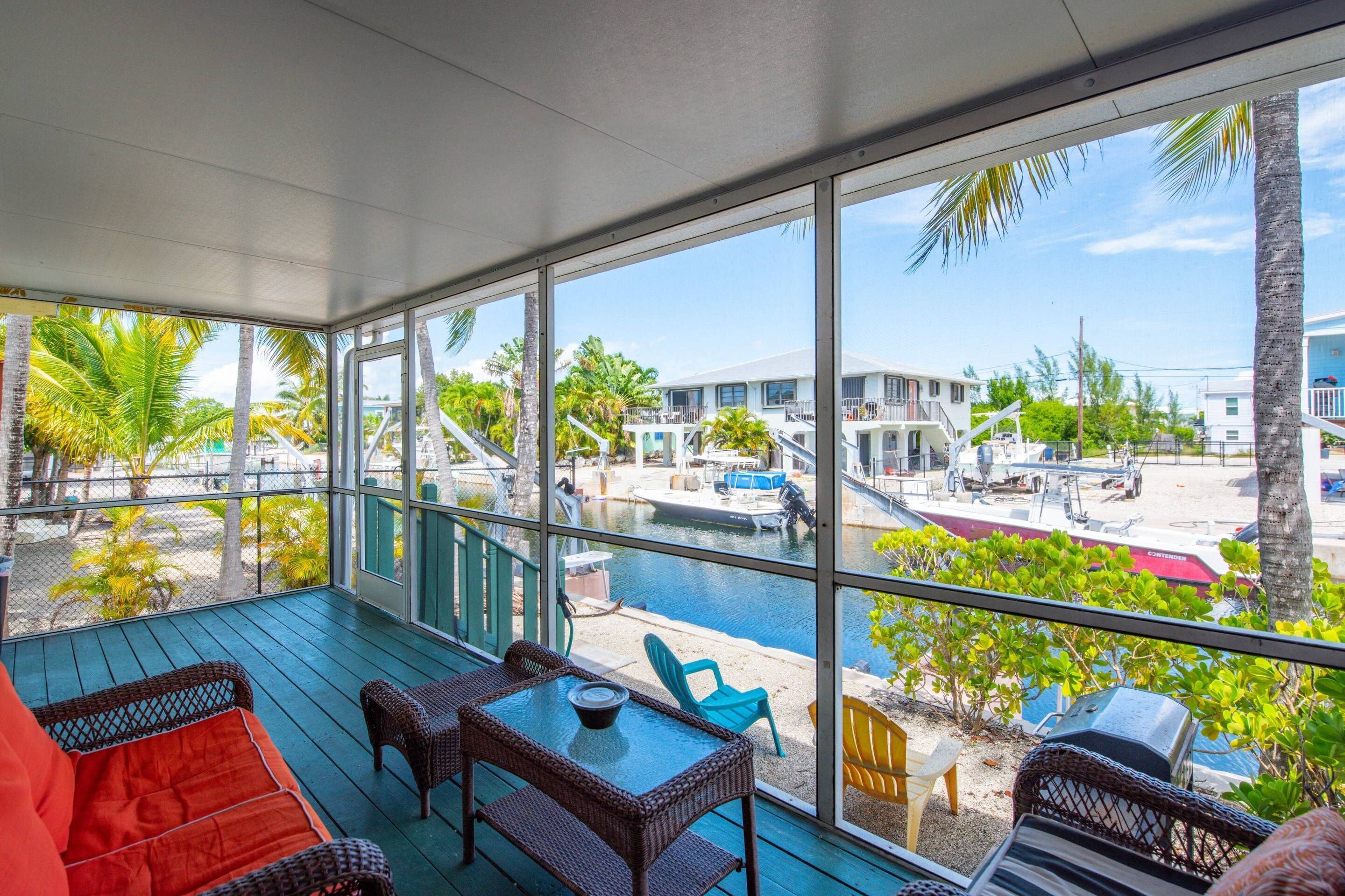 A covered outdoor area with wicker furniture, a glass table, and a view of a marina with boats and buildings.