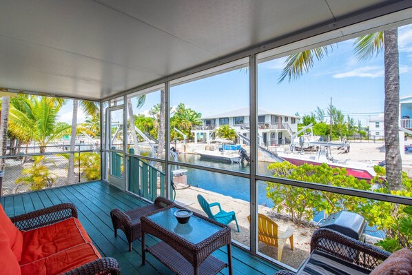 A covered outdoor area with wicker furniture, a glass table, and a view of a marina with boats and buildings.