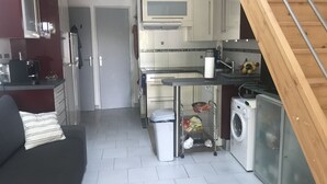 A compact kitchen with a microwave, stove, and washing machine. There is a small trash bin and a cart with cleaning supplies.