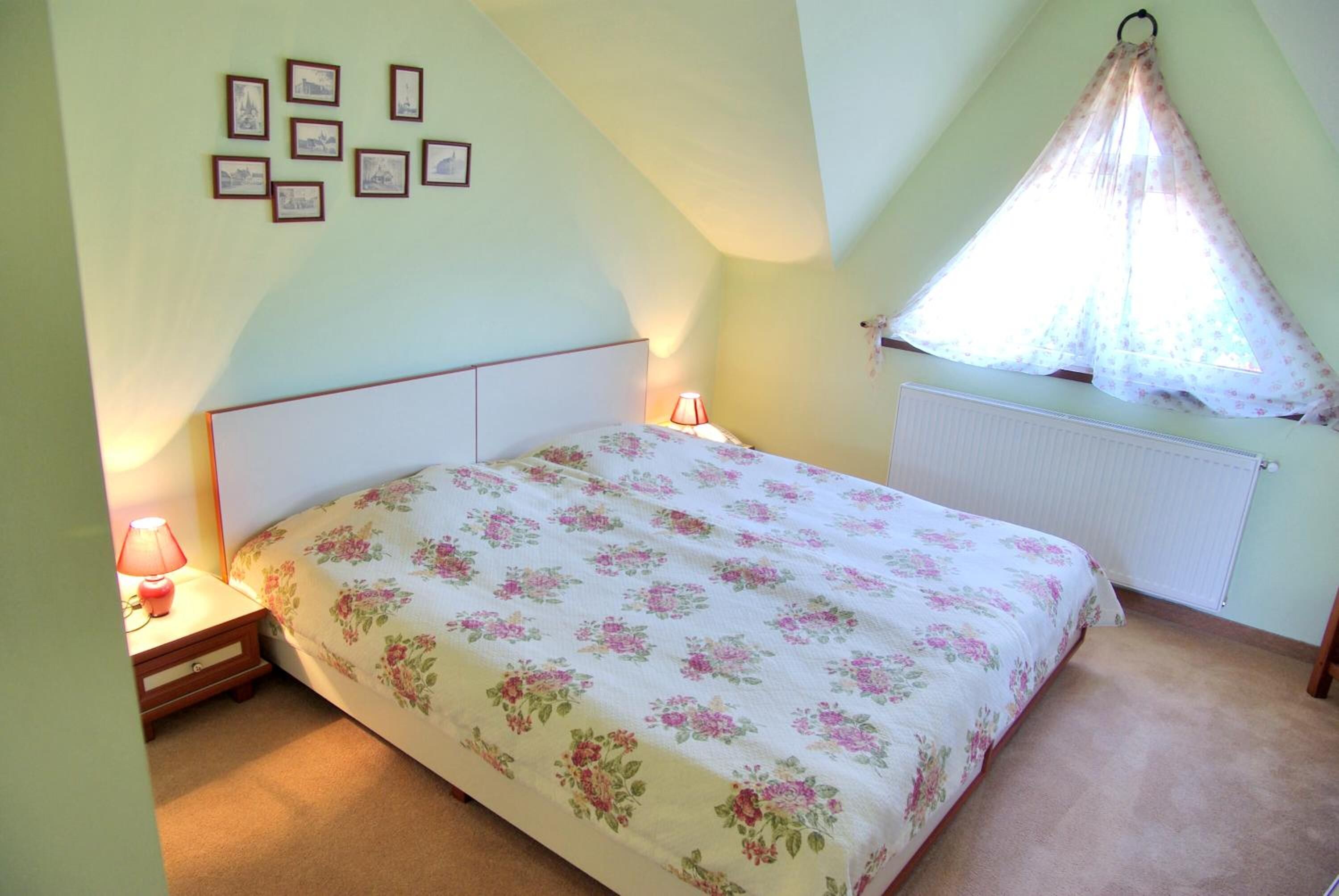 Un dormitorio con cama, mesita de noche, lámpara, ventana con cortina y cuadros en la pared.