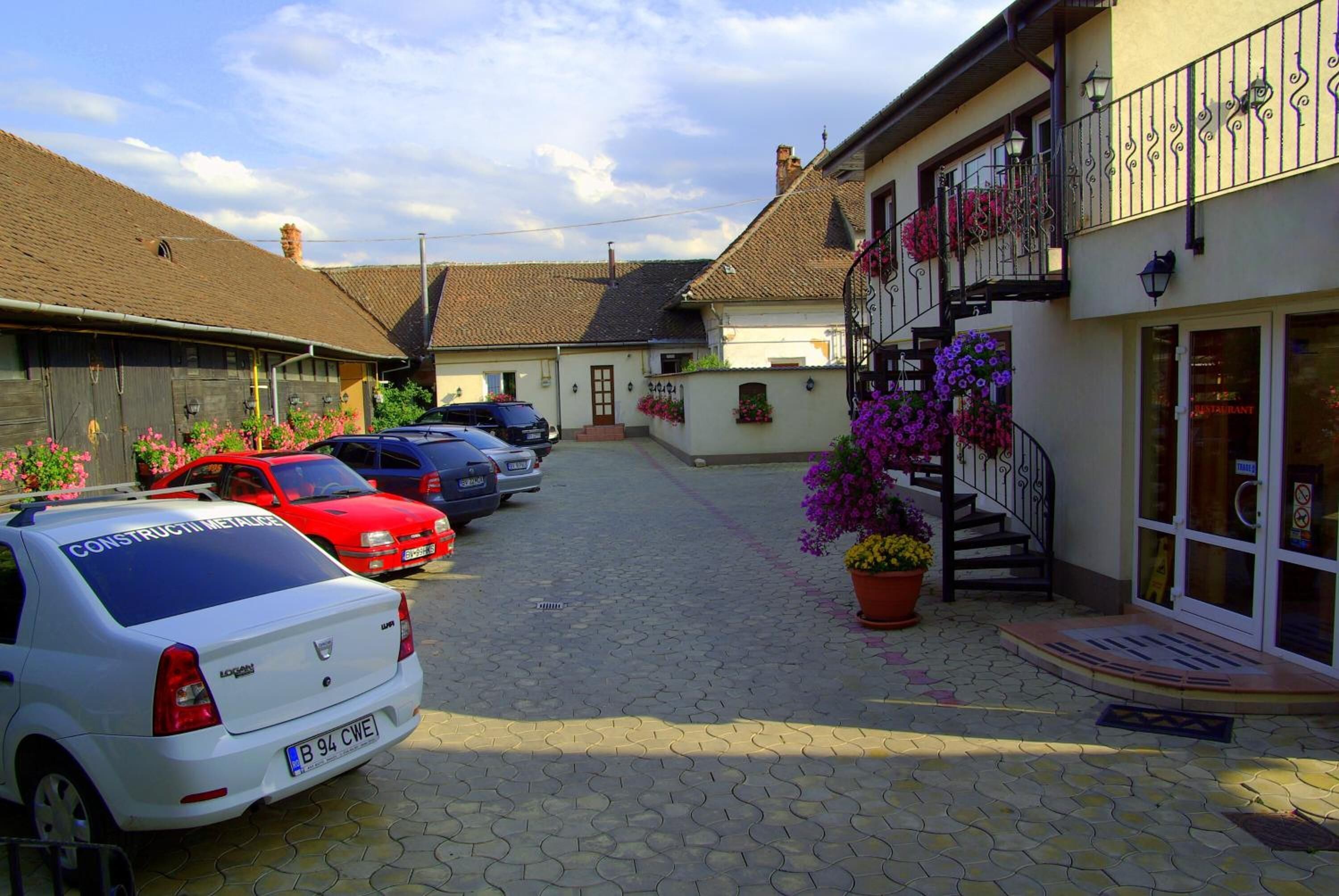 Una estrecha calle adoquinada con coches aparcados, un edificio con balcón decorado con flores y la entrada de un restaurante.