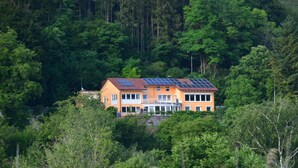 Une maison à deux étages, avec un toit rouge et des panneaux solaires, entourée d’une forêt dense.