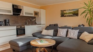 A modern living room with a grey sofa, a round wooden coffee table, and a kitchen area with white cabinets and appliances.