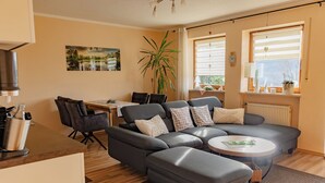 A modern living room with a grey sectional sofa, a dining area with a table and chairs, and large windows with blinds.