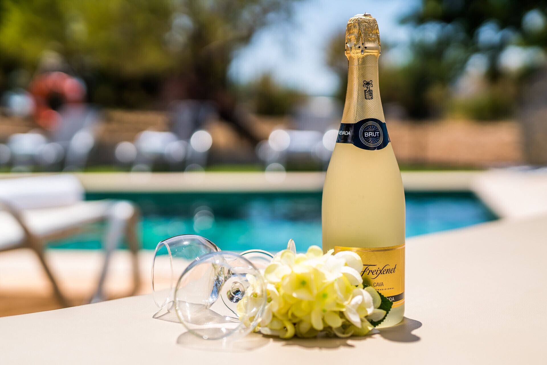 Una botella de champán Freixenet con un ramo de flores a su lado, sobre una mesa cerca de una piscina.