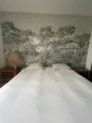 Une chambre à coucher comprenant un lit, deux oreillers, une table de chevet, une lampe et une grande tapisserie murale représentant un paysage.