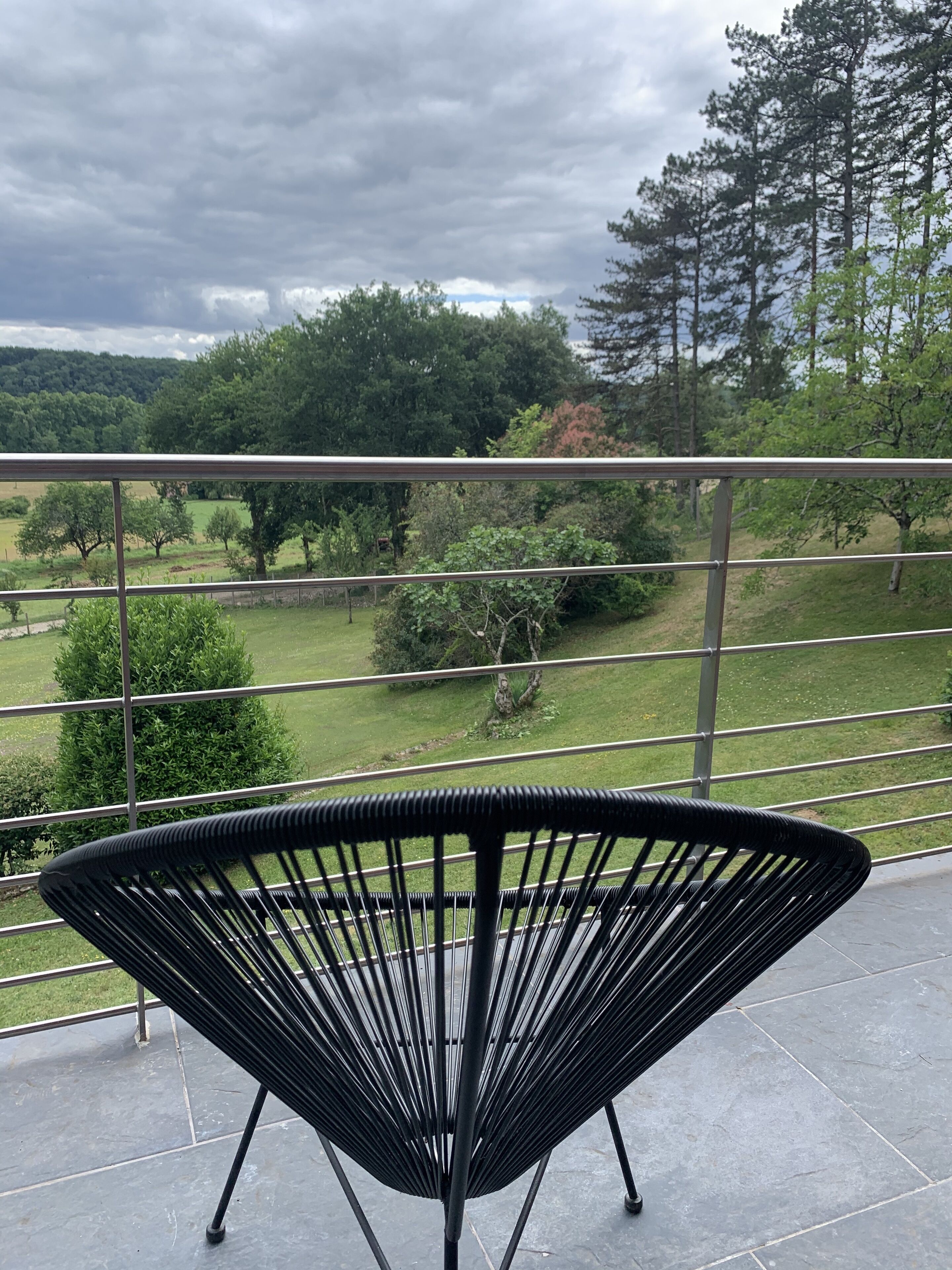 A balcony with a black chair, a metal railing, and a view of a green landscape with trees and a hill.