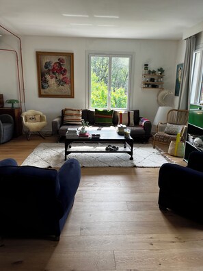 A living room with a sofa, armchairs, a coffee table, a painting, a window with a view of greenery, and a bookshelf.