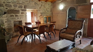 Een kamer met stenen muren, een houten eettafel met stoelen, een gestreept fauteuil en een betegelde vloer.