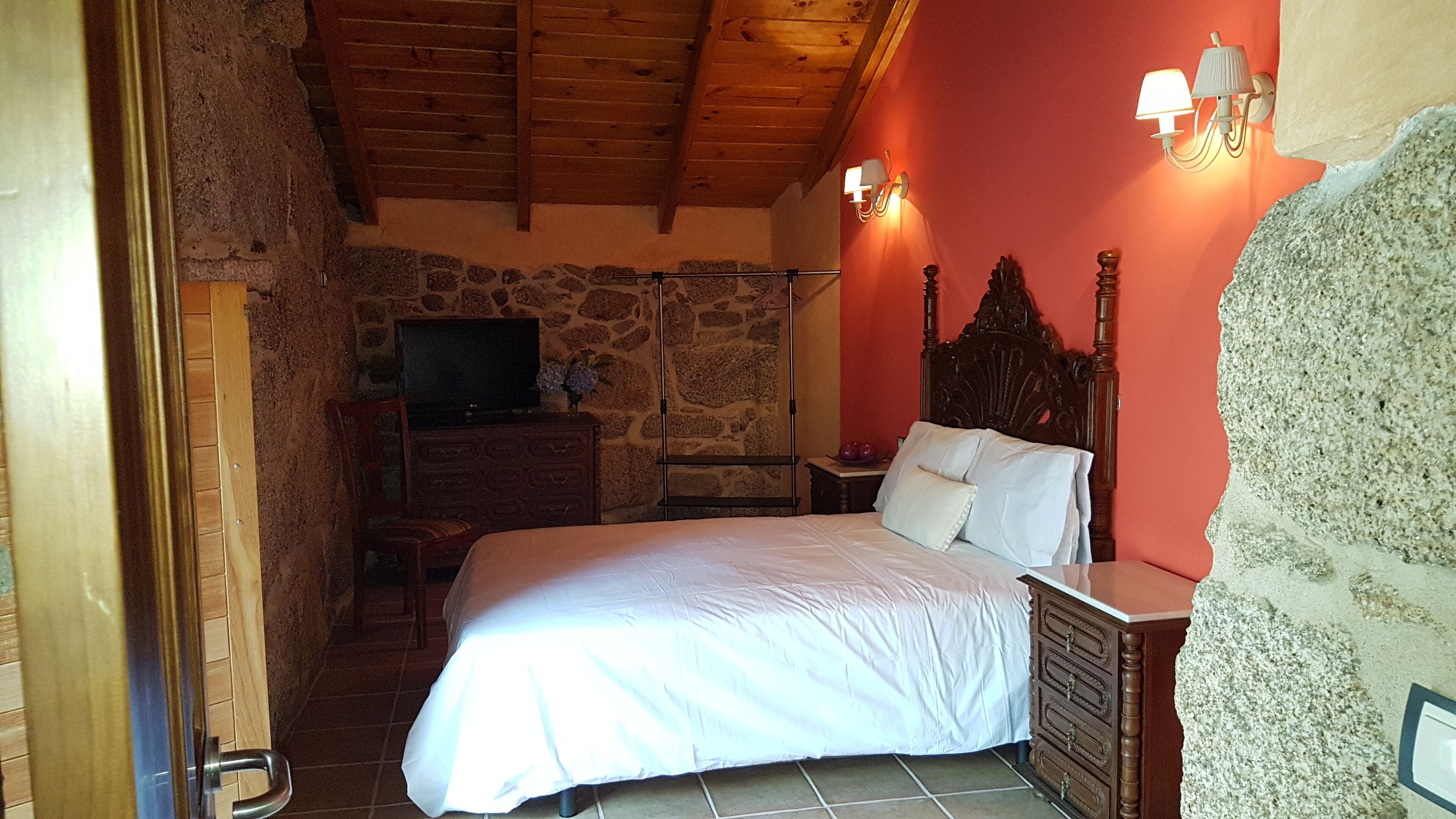 A bedroom with a stone wall, wooden ceiling, a bed with white linens, a wooden dresser, a TV on a stone shelf, and two wall-mounted lamps.