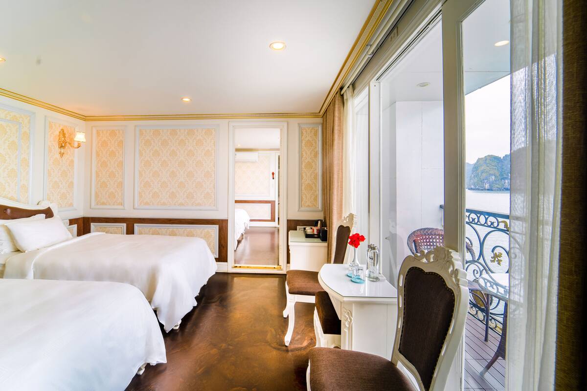 A hotel room with two beds, a small table with a vase of flowers, a chair, and a balcony with a view of the sea.