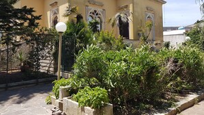 Un edificio con detalles arquitectónicos ornamentales, una entrada con reja y abundante vegetación en primer plano.