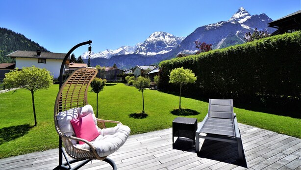 Une terrasse avec un fauteuil en osier et une chaise longue, une plateforme en bois et une vue sur des montagnes enneigées.