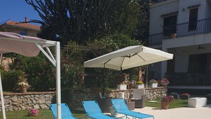 A poolside area with green lounge chairs, a white umbrella, and a stone wall with potted plants. - Villa Flore - Loft - (Casteldaccia)