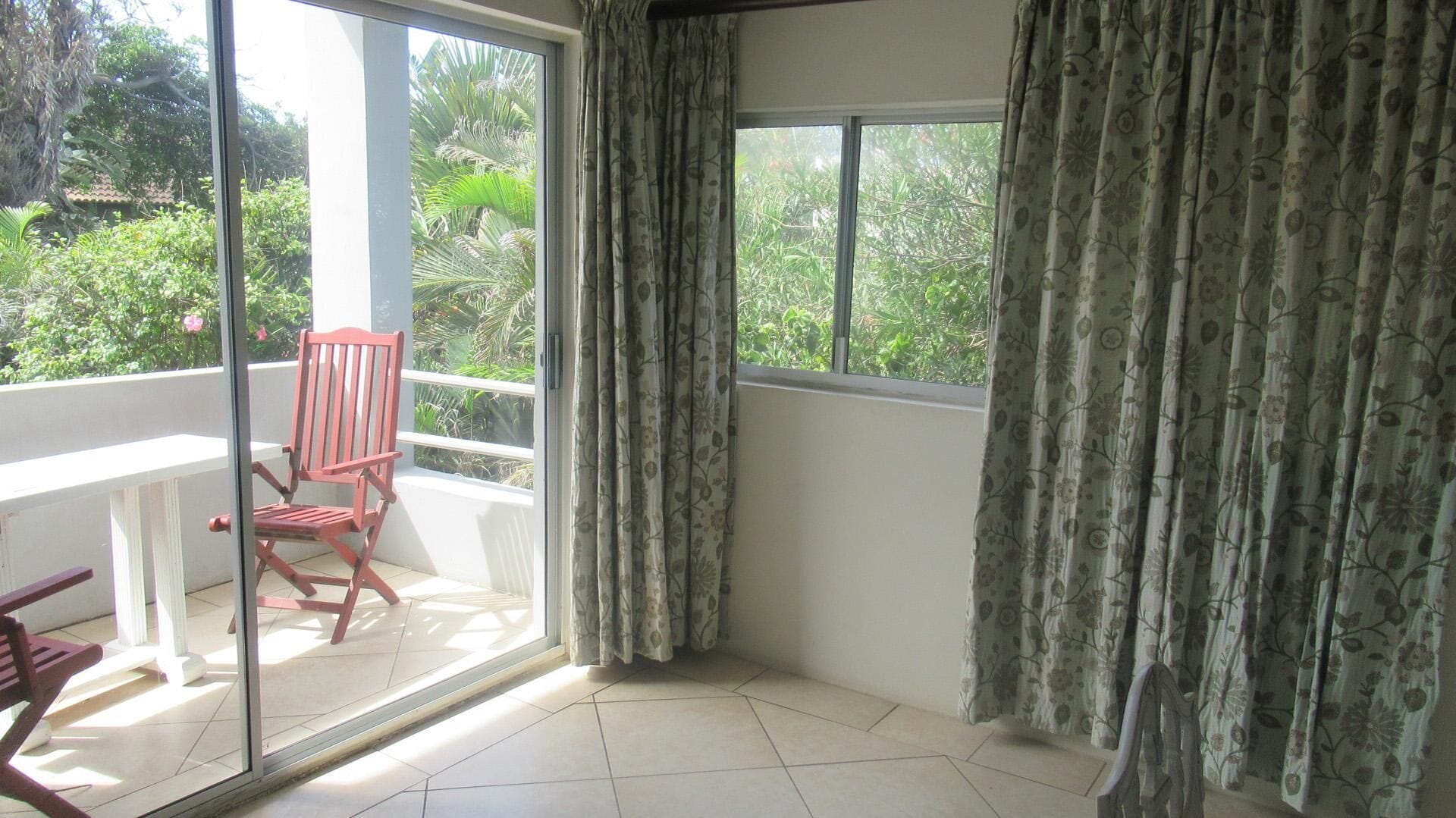 A balcony with a red chair, a white table, and a view of greenery through the window.