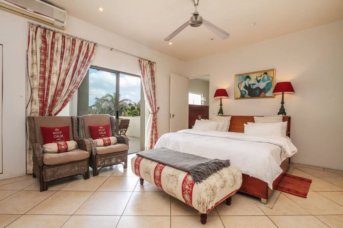 A bedroom with a bed, two chairs, a ceiling fan, a painting, and a window with curtains.