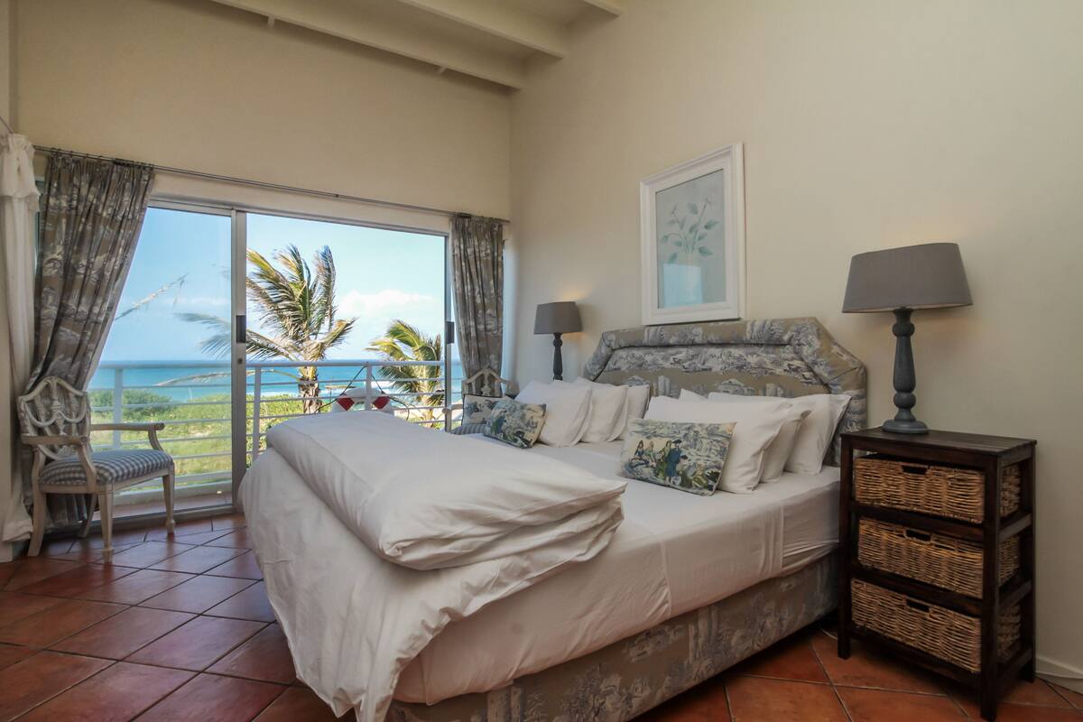 A bedroom with a large bed, a nightstand, a chair, and a view of the ocean.