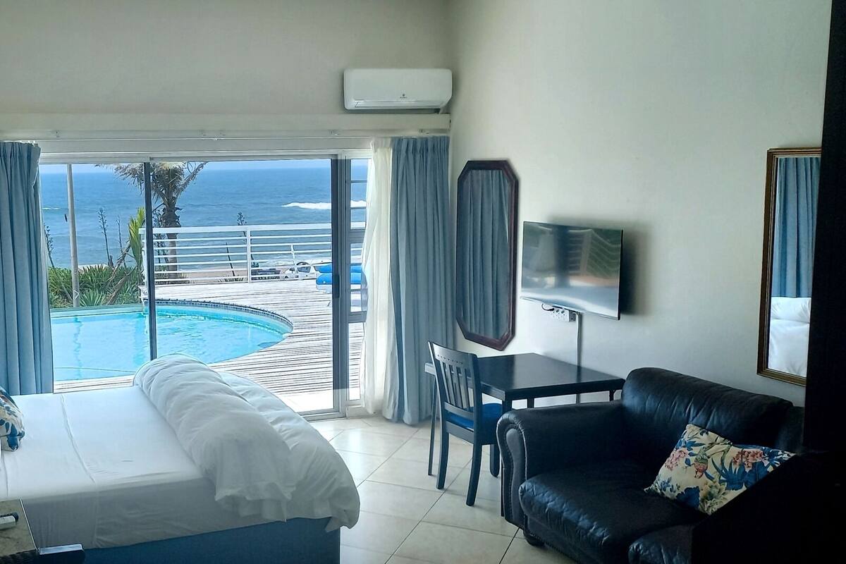 A hotel room with a bed, a desk, a chair, a TV, and a view of a pool and ocean.