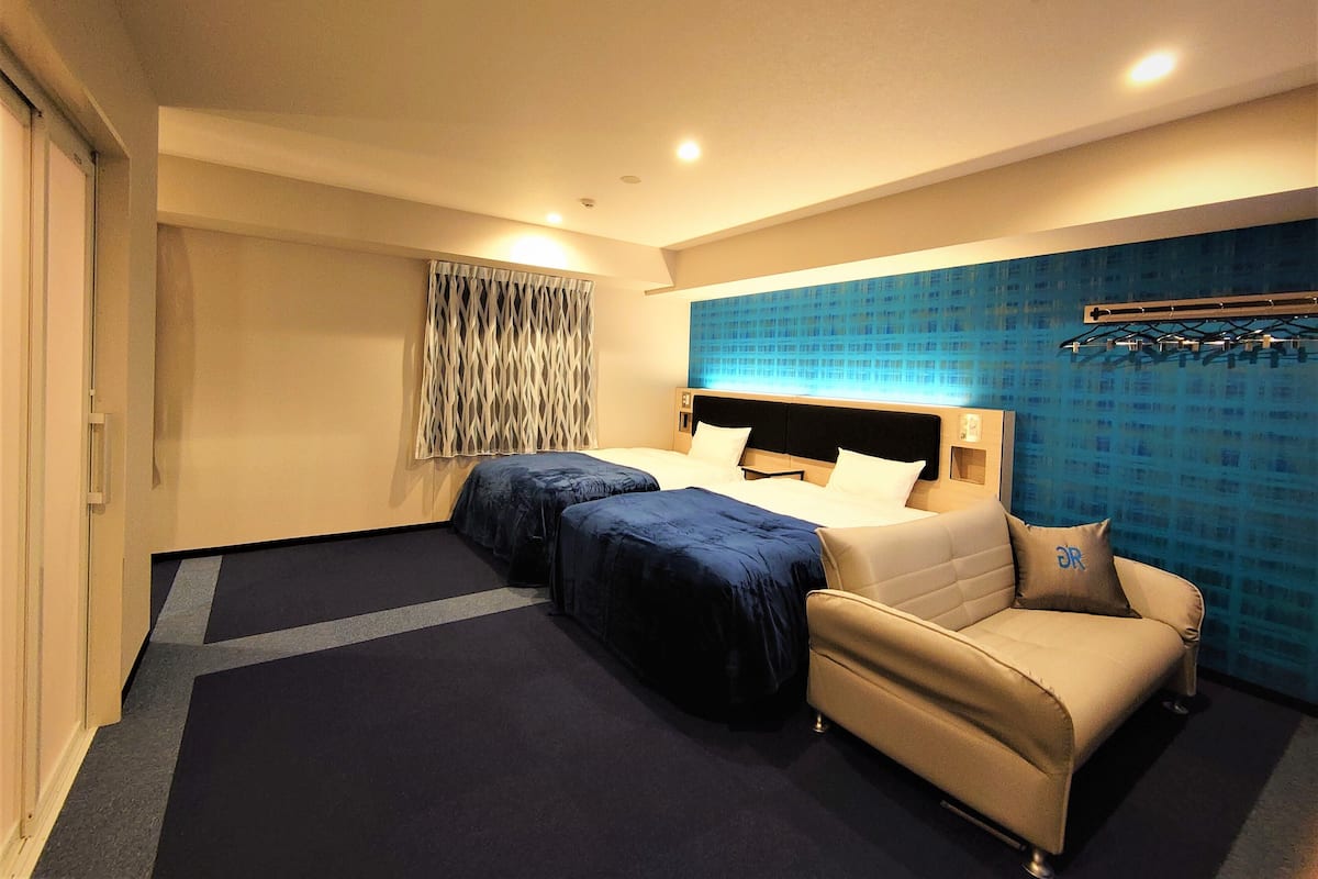 Habitació d'hotel amb un llit gran, dues taules de nit, un sofà, una finestra amb cortines i una paret blava amb un dibuix relleu.