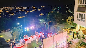 Evento al aire libre con mesas y sillas, rodeado de vegetación y edificios al fondo.