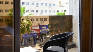 Een balkon met een stoel en een tafel, uitzicht op een gebouw met meerdere verdiepingen en reclameborden voor mobiele providers, en een heldere lucht.