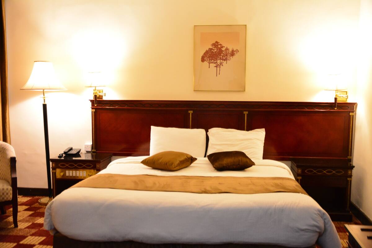 Una habitación de hotel con una cama grande, dos lámparas de noche, una mesita de noche con un teléfono y un cuadro en la pared.