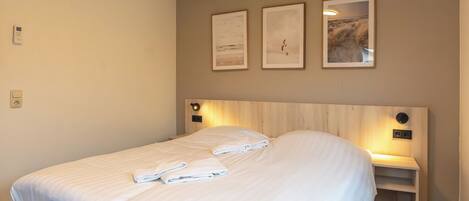 Un lit bien fait, avec du linge de lit et des serviettes blanches, une tête de lit en bois, des lampes de chevet et trois tableaux encadrés accrochés au mur.