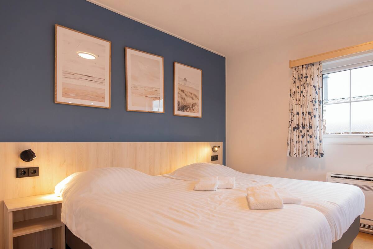 A bedroom with a large bed, a bedside table, a wall-mounted light, and a window with curtains.