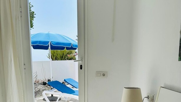Une chambre à coucher comprenant un lit, une table de chevet, une lampe et donnant sur un espace extérieur aménagé avec des parasols et des chaises longues.