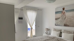Une chambre à coucher avec un lit, une table de chevet, un tableau représentant une scène de plage et une fenêtre avec des rideaux.