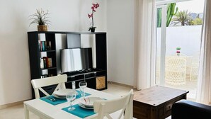 Un coin repas avec une table et des chaises blanches, une bibliothèque noire, un téléviseur et une vue sur un jardin de cactus, visible à travers une porte coulissante.